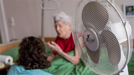 Eine Pflegerin hört einer pflegebedürftigen Seniorin zu. Ein Standventilator steht im Zimmer.