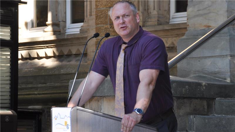 Ein Mann in lila Hemd und Krawatte steht an einem Rednerpult vor einem historischen Gebäude mit Steintreppe und hält sich an der Pultkante fest.