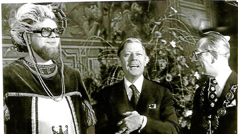 Drei Männer in formeller Kleidung stehen vor einer Wand mit dekorativen Elementen und Pflanzen im Hintergrund