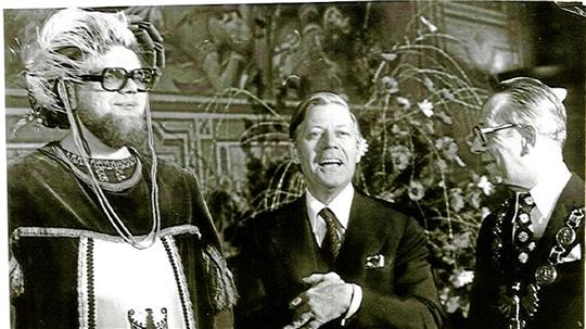 Drei Männer in formeller Kleidung stehen vor einer Wand mit dekorativen Elementen und Pflanzen im Hintergrund