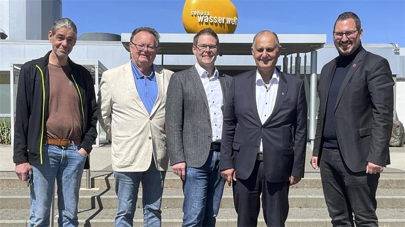 Fünf Männer in unterschiedlicher Kleidung stehen vor einer Treppe und einem Gebäude mit einer orangefarbenen Kugel und dem Schriftzug 'erlebnis wasserwelt'