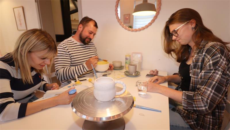 Drei Personen sitzen an einem Tisch. Auf dem Tisch stehen Teller, Gläser und eine weiße Keramik-Tasse.