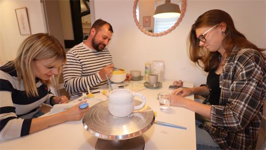 Drei Personen sitzen an einem Tisch. Auf dem Tisch stehen Teller, Gläser und eine weiße Keramik-Tasse.