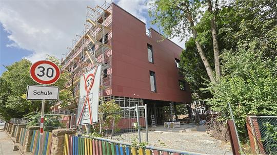 Modernes mehrstöckiges Schulgebäude mit roter Fassade und Baustellengerüst an der Seite, davor bunte Holzzäune und Verkehrsschilder mit Geschwindigkeitsbegrenzung 30 und dem Zusatz "Schule"