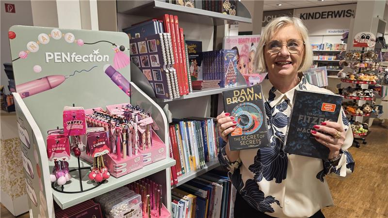 Person steht in einer Buchhandlung und hält zwei Bücher, eines davon von Dan Brown, vor einem Regal mit Kinder- und Jugendbüchern.