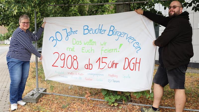 Zwei Personen halten ein großes weißes Banner mit bunter Schrift, das eine Einladung zur Feier des 30-jährigen Bestehens des Beuchter Bürgervereins zeigt