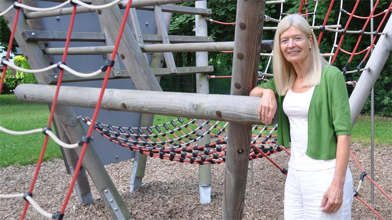 Frau mit grüner Jacke lehnt an einem Klettergerüst aus Holz und Seilen auf einem Spielplatz.