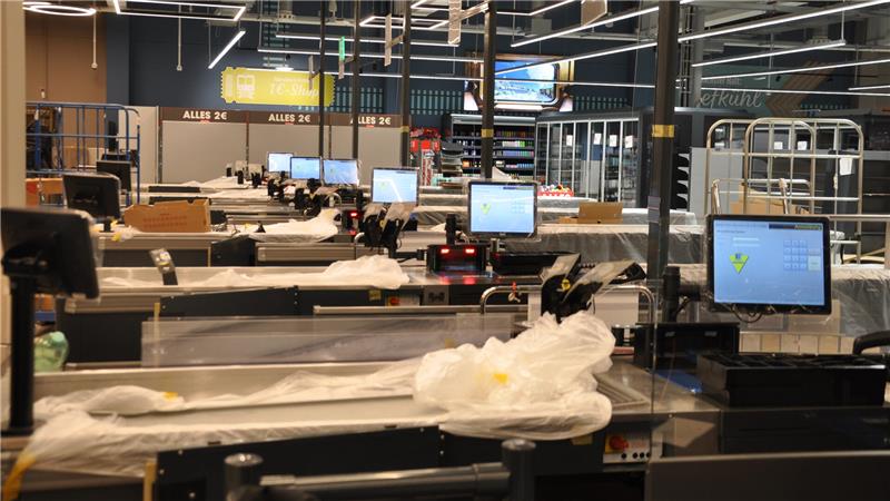 Leere Supermarktkassen mit Monitoren und Plastikabdeckungen, Deckenbeleuchtung und Regale im Hintergrund.