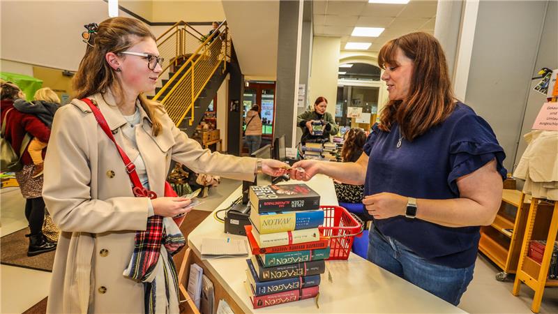 GZ-Volontärin Hanna Schlimme steht links im Bild und reicht Bibliotheksleiterin Claudia Holtkamp Geld über den Tresen. Im Vordergrund ist ein großer Stapel Bücher zu sehen. 