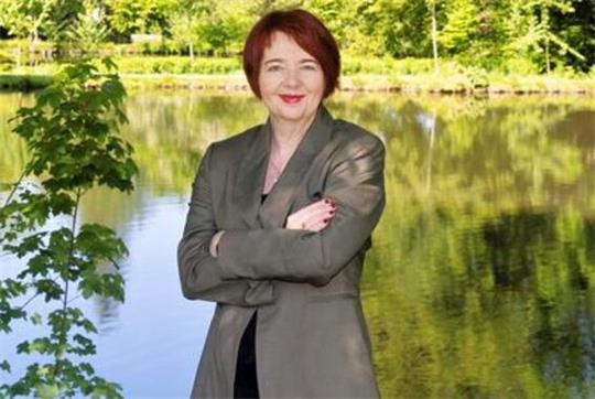 Frau mit rotem Haar und grauem Blazer steht mit verschränkten Armen vor einem See und grünen Bäumen im Hintergrund