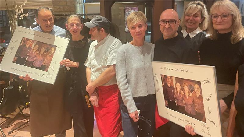 Gruppe von sieben Personen, zwei halten gerahmte Bilder mit mehreren Personen darauf, eine trägt Kochkleidung mit roter Schürze.