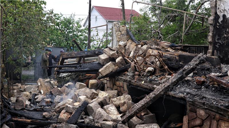 Trümmer eines zerstörten Hauses mit rauchenden Steinen und verkohltem Holz, im Hintergrund ein Mann vor einem Tor und ein Haus mit rotem Dach.