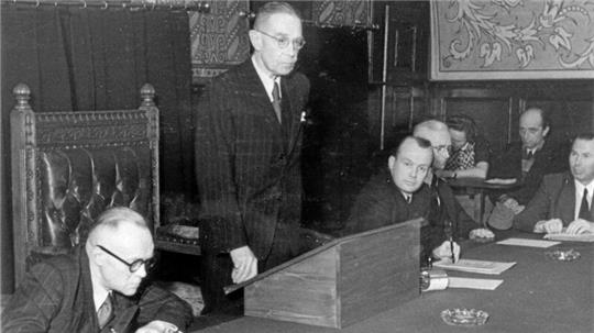 Mann in Anzug steht an einem hölzernen Rednerpult in einem Raum mit mehreren sitzenden Personen und verzierten Wänden.