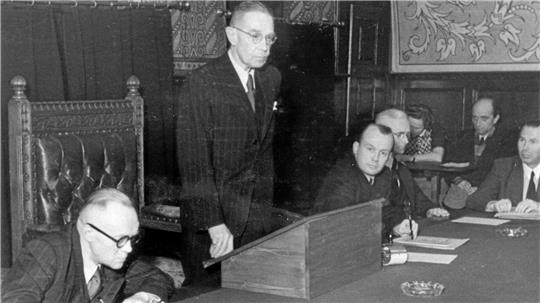 Mann in Anzug steht an einem hölzernen Rednerpult in einem Raum mit mehreren sitzenden Personen und verzierten Wänden.