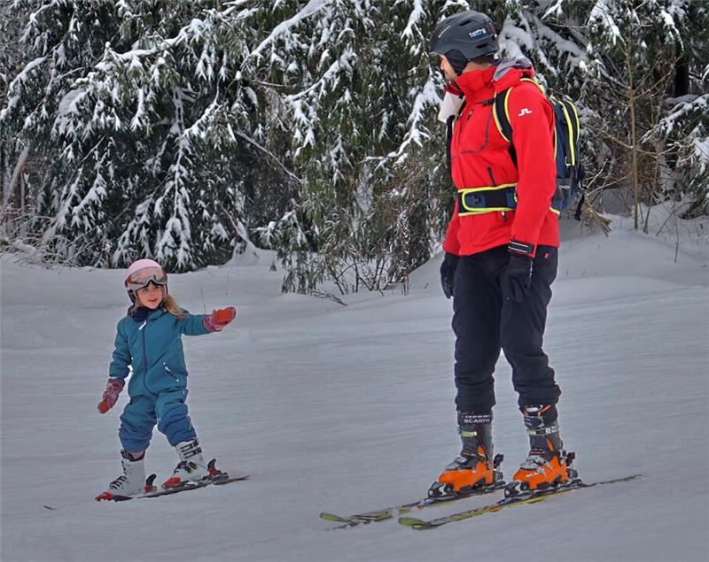 Kind in blauer Skibekleidung steht auf Skiern und streckt einen Arm aus, vor einem Erwachsenen in roter Skijacke und Helm auf Skiern im Schnee vor verschneiten Tannenbäumen.