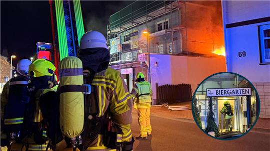 Feuerwehrleute mit Atemschutzgeräten stehen nachts vor einem brennenden Gebäude, während Flammen und Rauch aus dem Dach schlagen; ein Feuerwehrmann betritt einen Biergarten-Eingang.