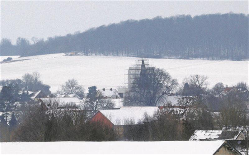 In schneebedeckter Landschaft ragt ein Kirchturm, den ein gerüst umgibt, aus einem Dorf. Im Hintergrund auf einem Bergrücken ein Wald.