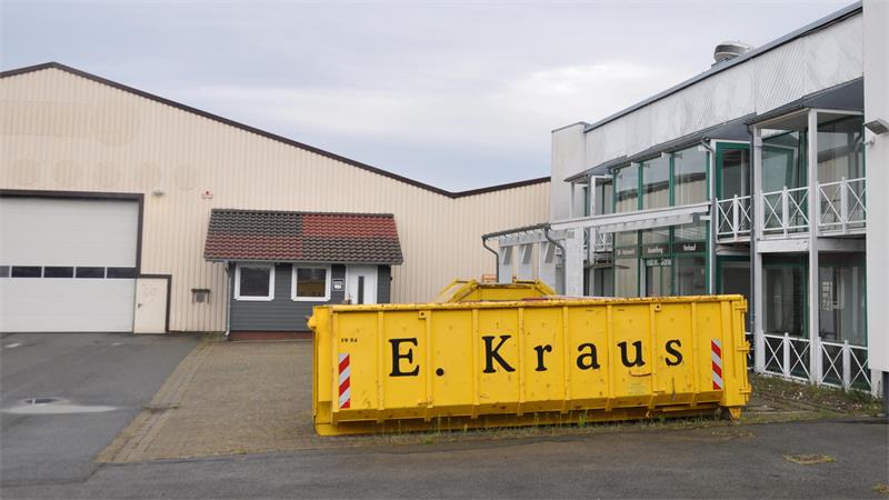 Gelber Abfallcontainer mit der Aufschrift 'E. Kraus' vor einem Industriegebäude mit Glasfassade und Lagerhalle