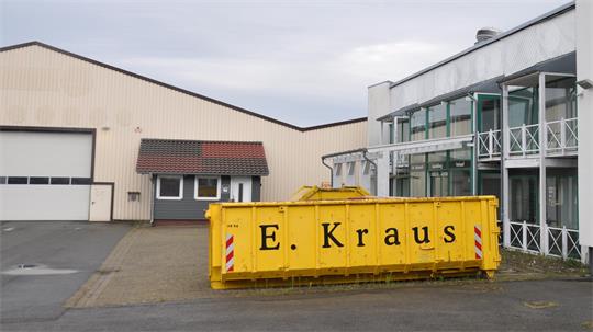 Gelber Abfallcontainer mit der Aufschrift 'E. Kraus' vor einem Industriegebäude mit Glasfassade und Lagerhalle