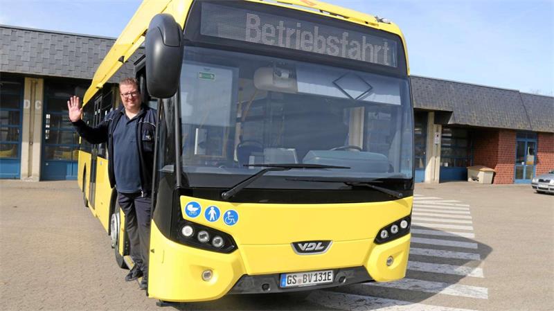 Mann winkt aus der offenen Tür eines gelben Busses mit der Aufschrift 'Betriebsfahrt'