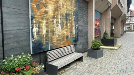 Holzbank vor einer Wand mit abstraktem Gemälde in einem urbanen Bereich mit Pflanzkästen und Kopfsteinpflaster