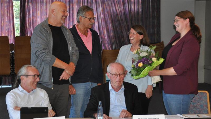 Sechs Personen in einem Raum, zwei sitzen an einem Tisch mit Namensschildern, vier stehen, eine Frau überreicht einer anderen Frau einen Blumenstrauß, alle lächeln.