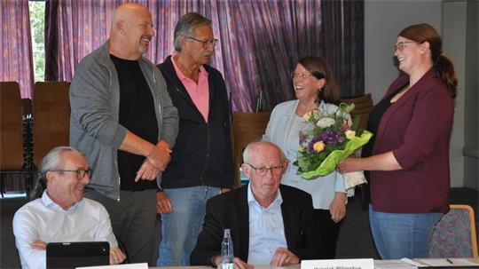 Sechs Personen in einem Raum, zwei sitzen an einem Tisch mit Namensschildern, vier stehen, eine Frau überreicht einer anderen Frau einen Blumenstrauß, alle lächeln.
