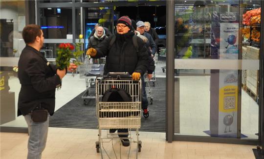 Mann mit schwarzer Jacke und roter Mütze schiebt Einkaufswagen durch automatische Glastür, weitere Personen im Hintergrund.