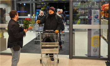 Mann mit schwarzer Jacke und roter Mütze schiebt Einkaufswagen durch automatische Glastür, weitere Personen im Hintergrund.