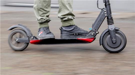 Person mit grauen Sneakers und grünen Hosen steht auf einem schwarzen und roten E-Scooter.