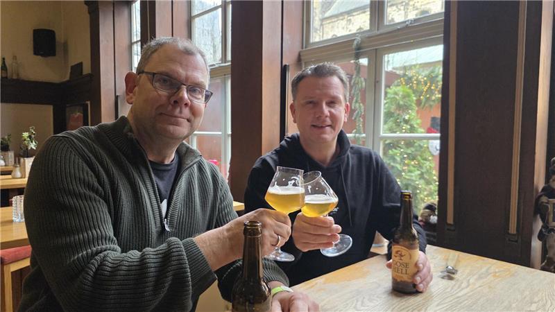 Zwei Männer sitzen an einem Holztisch in einem Gastraum und stoßen mit Biergläsern an, auf dem Tisch stehen zwei Bierflaschen.