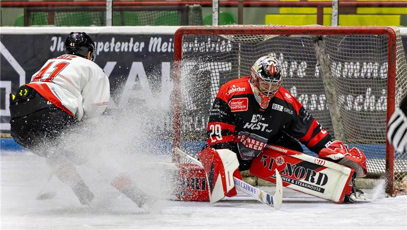 Eishockeyspieler in weißem Trikot schießt auf das Tor, Torwart in schwarzem und rotem Schutzanzug hält den Puck nicht.
