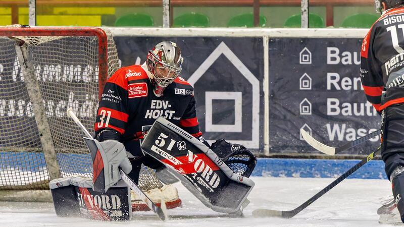 Eishockeytorwart in schwarzer und roter Ausrüstung hält den Puck vor dem Tor auf dem Eis fest