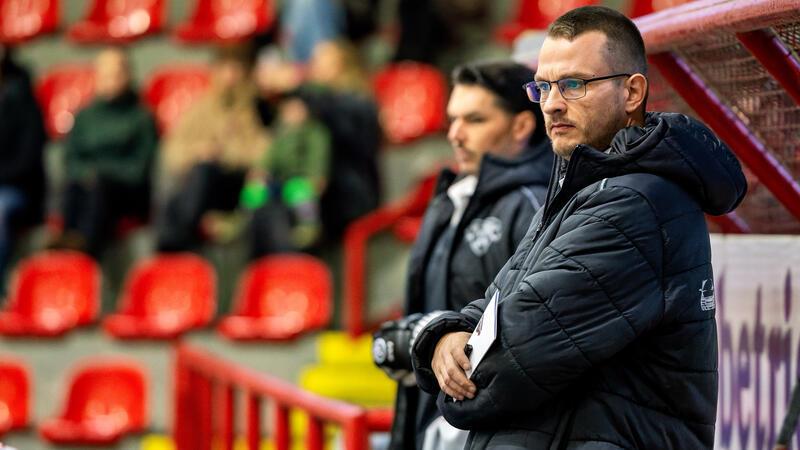 Mann mit Brille und schwarzer Jacke lehnt mit verschränkten Armen an einer Bande in einer Sporthalle mit roten Sitzen
