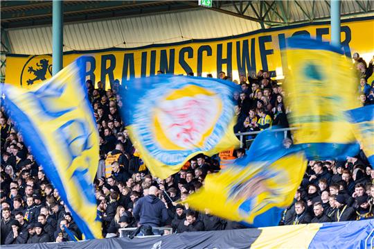 Braunschweiger Fans stehen auf einer Tribüne im Stadion und schwenken mit verschiedenen Fahnen. Foto: picture alliance/dpa | Michael Matthey