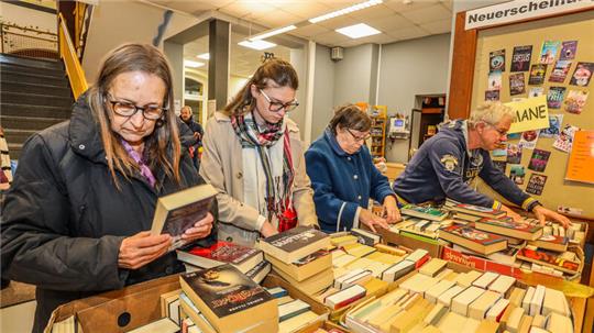 Vier Menschen stöbern in Kisten voller Bücher. Die Frau ganz links hält ein Buch in beiden Händen.