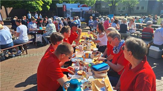 Menschen sitzen an langen Tischen im Freien und essen gemeinsam bei Sonnenschein