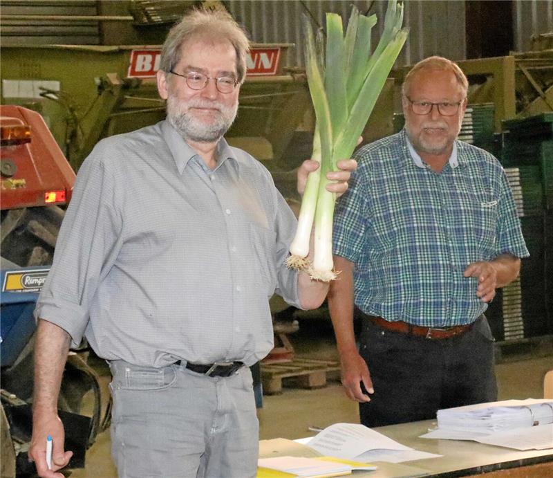 Zwei Männer stehen in einer Halle, einer hält mehrere Lauchstangen in der Hand, auf dem Tisch liegen Aktenordner und Papiere.