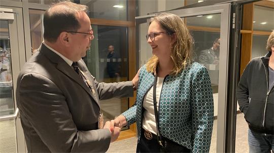 Zwei Personen in Innenraum schütteln sich lächelnd die Hand, eine trägt Anzug, die andere eine gemusterte Jacke