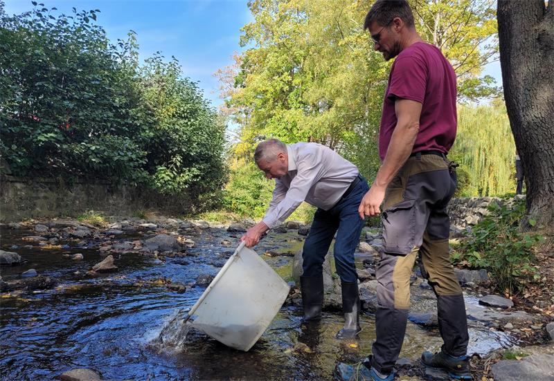 Zwei Männer an einem Fluss. Einer kippt einen großen weißen Bottich aus. 
