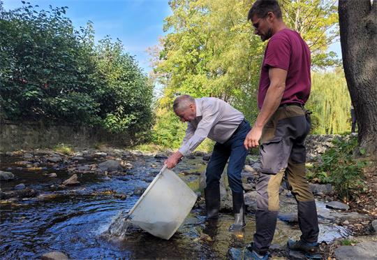 Zwei Männer an einem Fluss. Einer kippt einen großen weißen Bottich aus. 