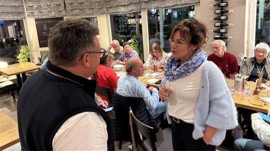 Das Foto zeigt eine Frau und einen Mann, die sich gegenüberstehen, und im Hintergrund einen voll besetzten Tisch.
