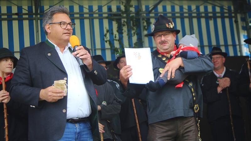 Bürgermeister Wolfgang Langer (links) ernennt Andreas Neigenfindt, der seinen fünf Monate alten Neffen Jano im Arm hält, zum Gemeindehirten.