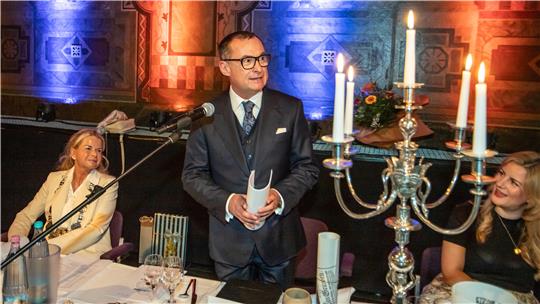 Mann in dunklem Anzug steht an einem Mikrofon vor einem Tisch mit Kerzenleuchter und mehreren Personen in einem festlich beleuchteten Raum