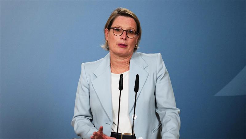 Frau mit Brille und hellblauem Blazer spricht vor zwei Mikrofonen vor blauem Hintergrund