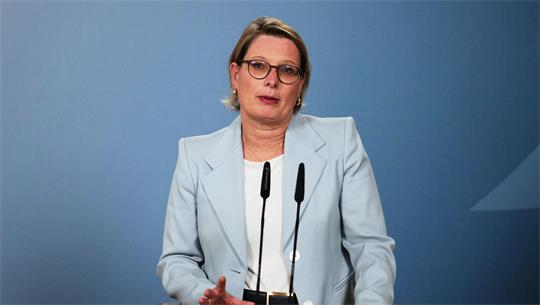 Frau mit Brille und hellblauem Blazer spricht vor zwei Mikrofonen vor blauem Hintergrund