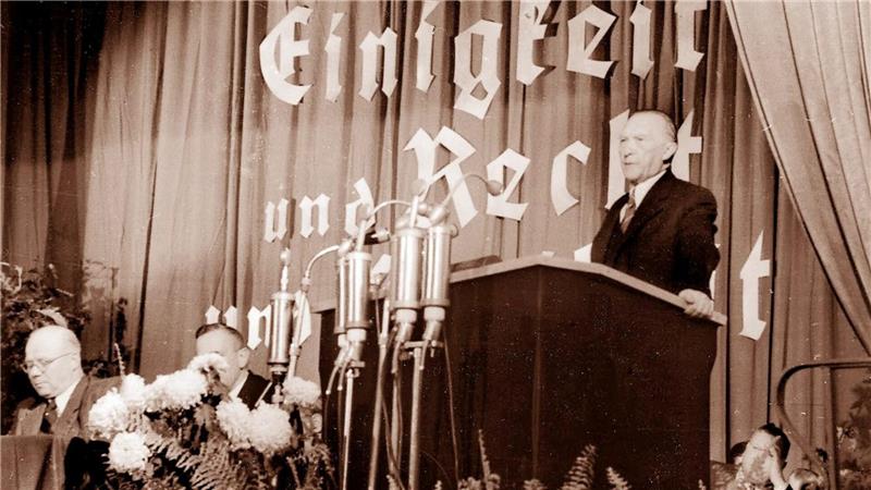 Mann steht an Rednerpult vor Vorhang mit großem Schriftzug und mehreren Mikrofonen