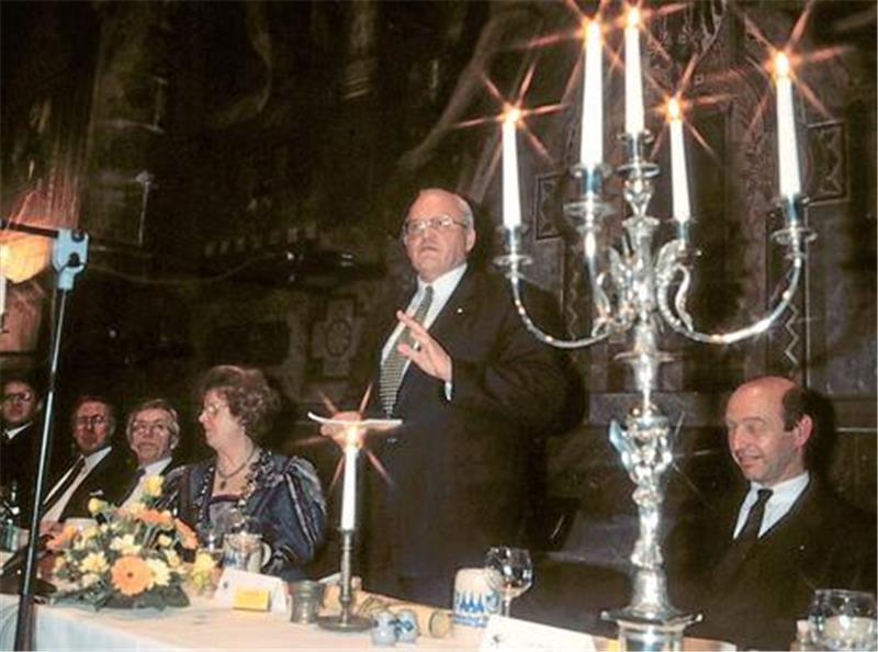 Mehrere Personen sitzen an einem festlich gedeckten Tisch mit Kerzenleuchtern und Blumenarrangements in einem dunklen Raum