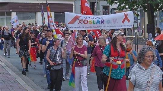 Menschenmenge bei einem Straßenumzug mit einem Banner der AIDSHILFE GOSLAR und mehreren Regenbogenfahnen