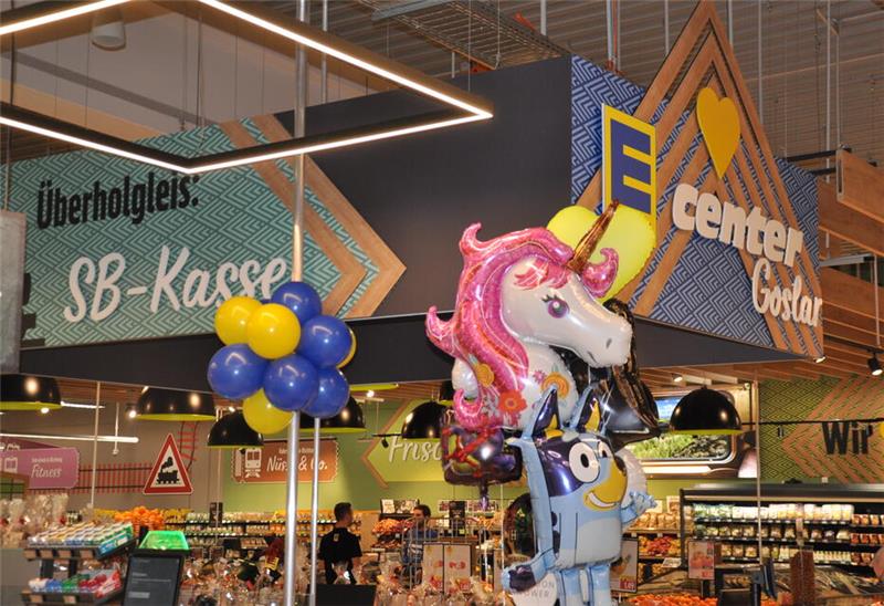 Goslarer Edeka-Center eröffnet: Erste Kunden kommen um sieben Uhr Bunte Luftballons in Form eines Einhorns und einer Figur mit Brille hängen in einem Supermarkt unter einem Schild mit der Aufschrift 'Überholgleis: SB-Kasse'.
